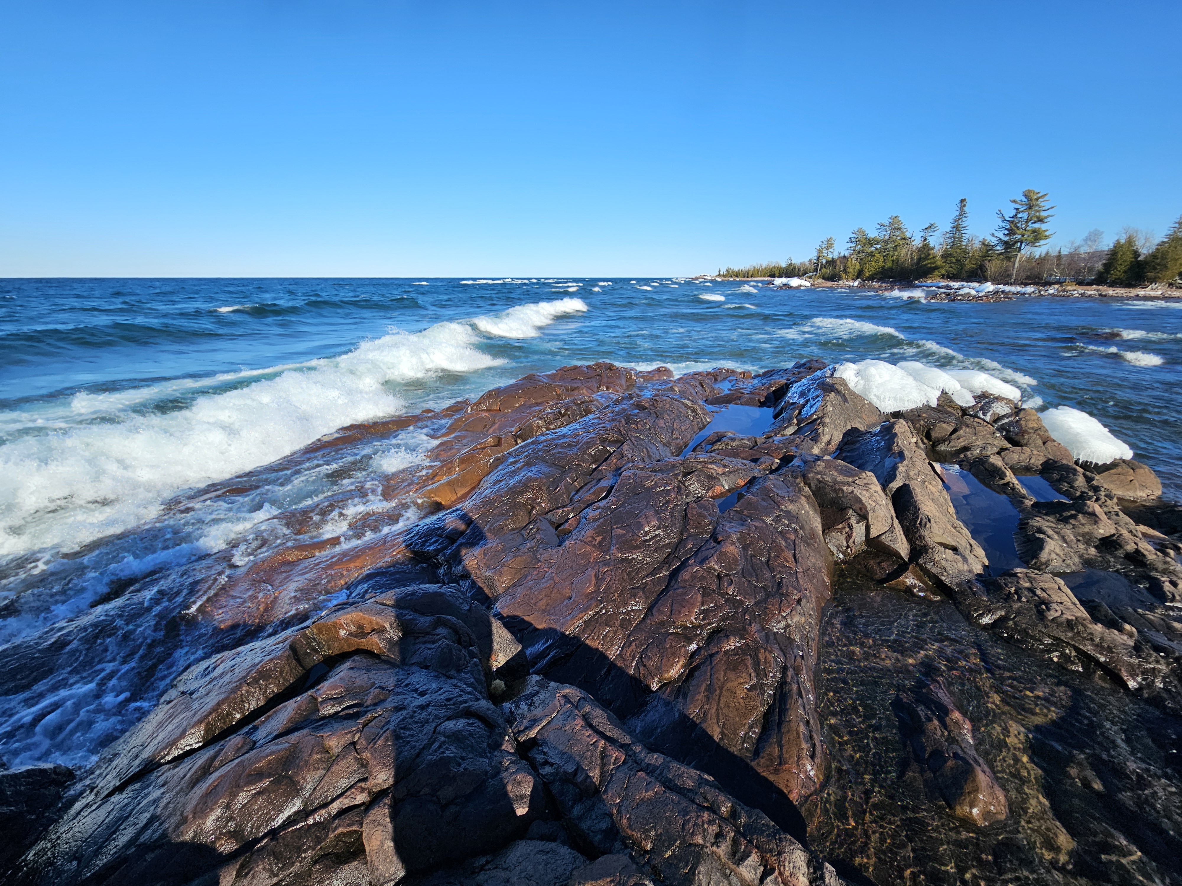 Emily's shadow stretches out across the ice- and snow-covered rocks of a stony outcrop into a turbulent and very blue Lake Superior.