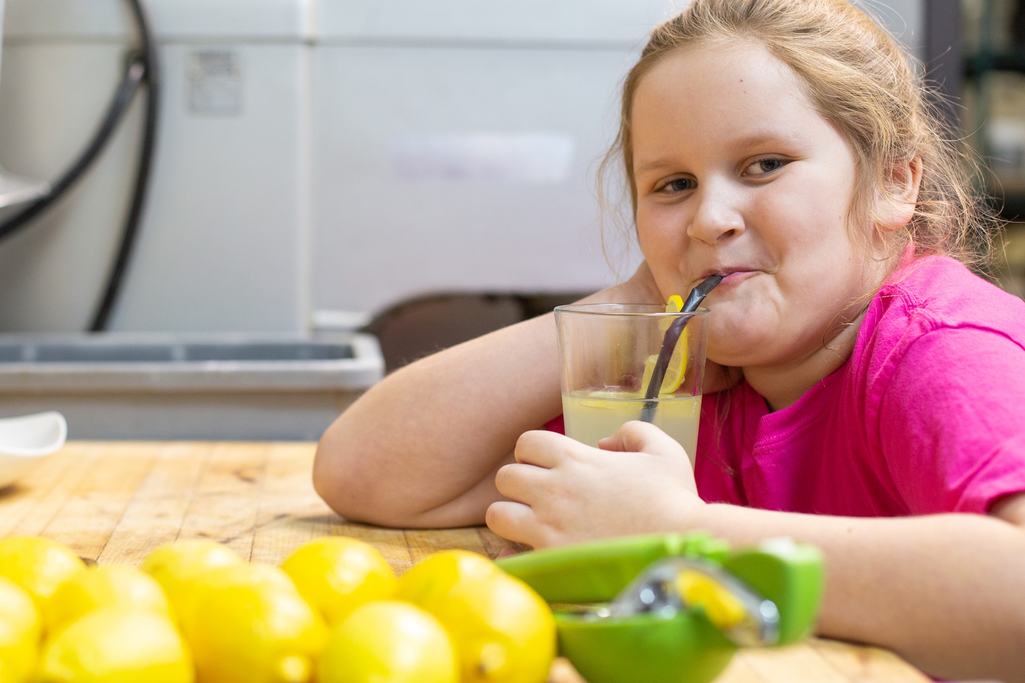 A young girl wearing a bright pink shirt sits at a table, smiling as she sips lemonade through a straw. Several lemons can be seen on the table in front of her, along with a juicing tool.