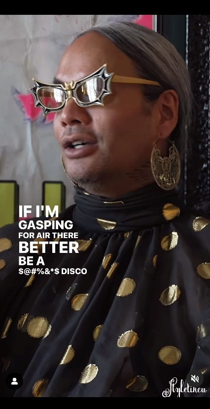 Screen grab showing Raja wearing glasses shaped like a bat with spiky wings, large and ornate gold earrings, a flowy black dress with gold polka dots, and her silver-gray hair pulled back in a pony tail.