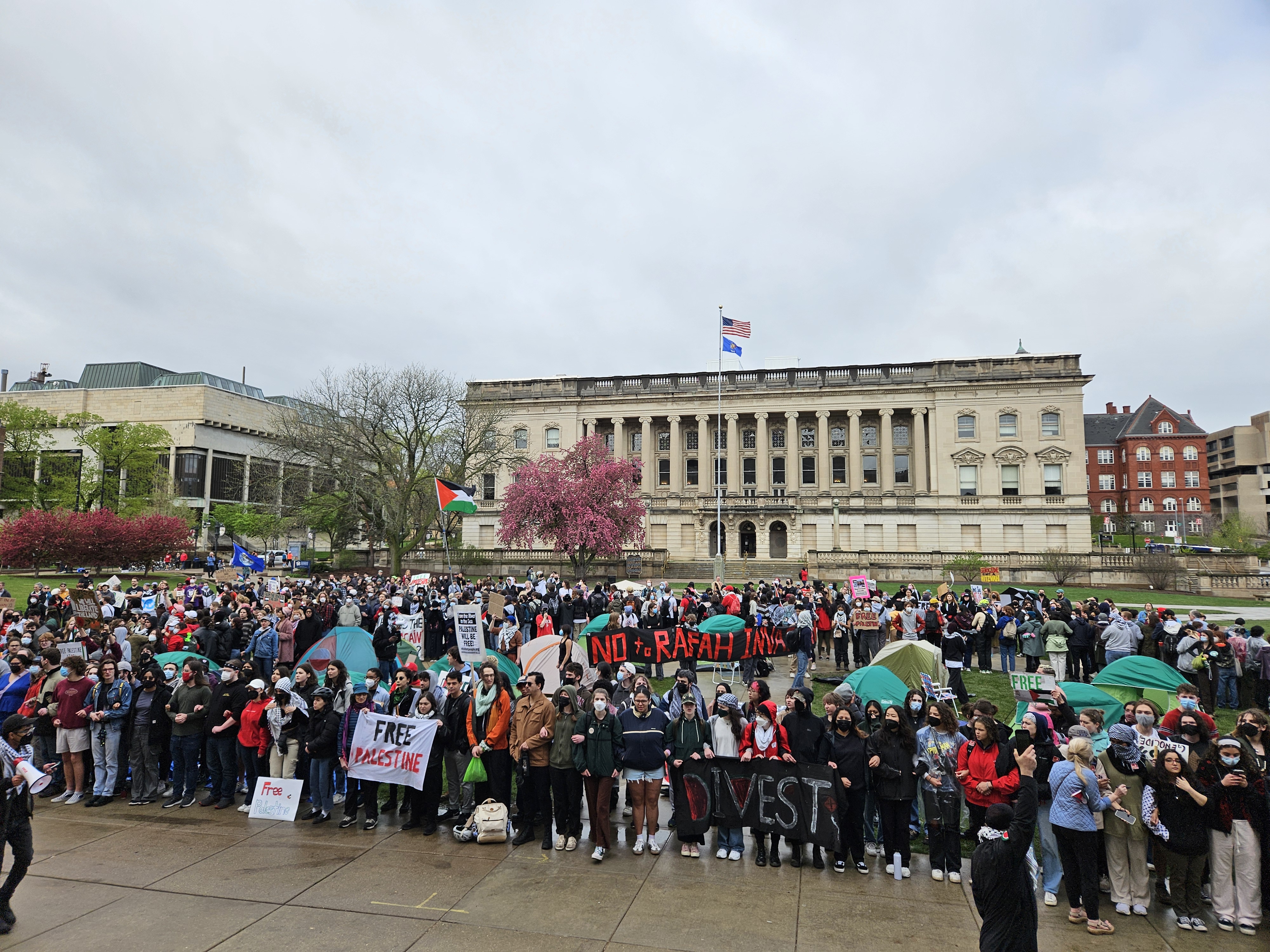 A large crowd of people stands in a circle around a cluster of people erecting camping tents. The scene is unfolding int he middle of a wide open campus quad, with school buildings in the background. A Palestinian flag waves from a pole at the center of the encampment, and the people hold various signs protesting the genocide in Gaza and demanding that the school divest from any funds that support that violence.