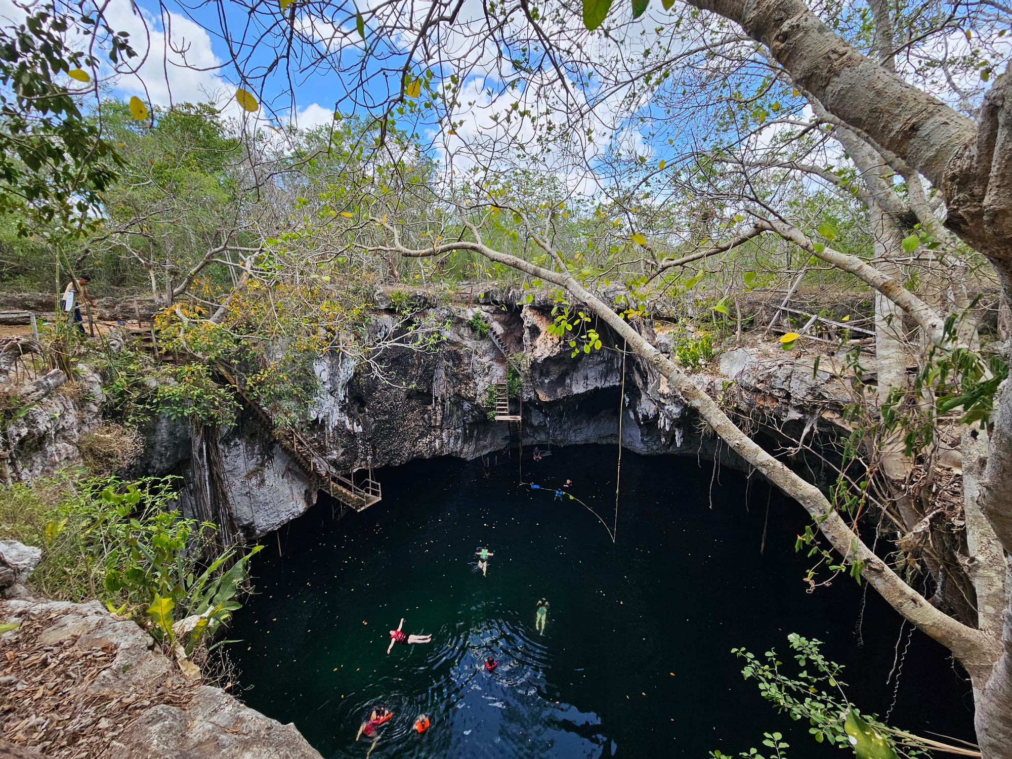 A large hole in the ground, surrounded by deep limestone walls and green forest, is filled with dark blue water where several people can be seen swimming.