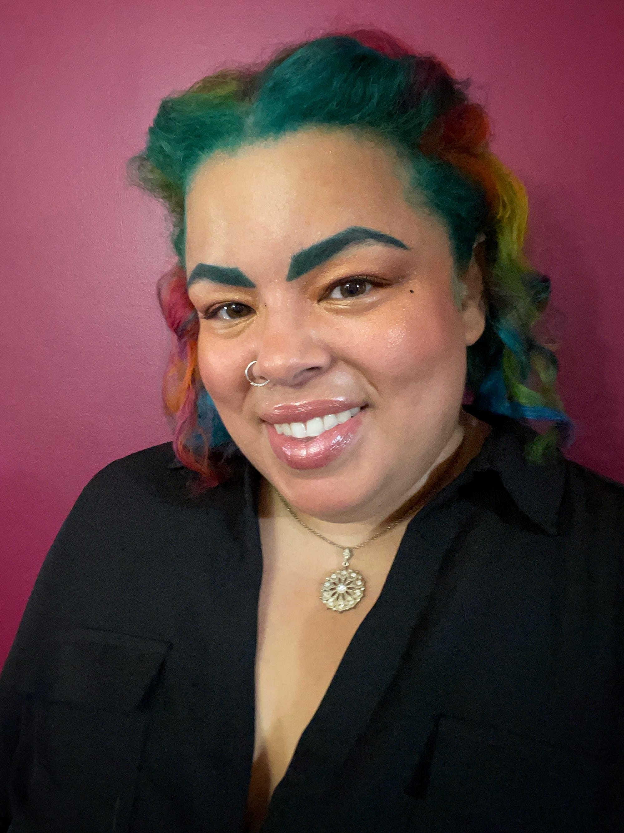 Valentine’s head shot. They are a ilght-skinned Black non-binary person with curly hair dyed rainbow colors and a nose piercing, and they are smiling.