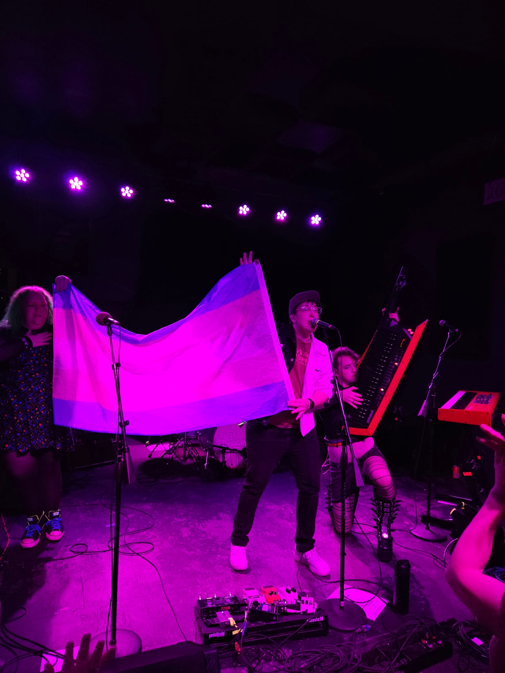 Three people stand on a stage, two of them holding the Transgender Pride Flag, and the other on their knees and playing a very large keytar.