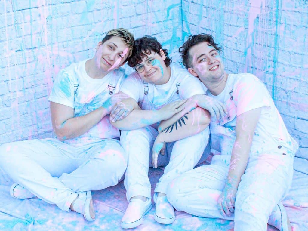 Three white non-binary humans sit on the floor and cuddle. They are all dressed in white overalls with white t-shirts, and covered in pink, and baby blue paint splatters.