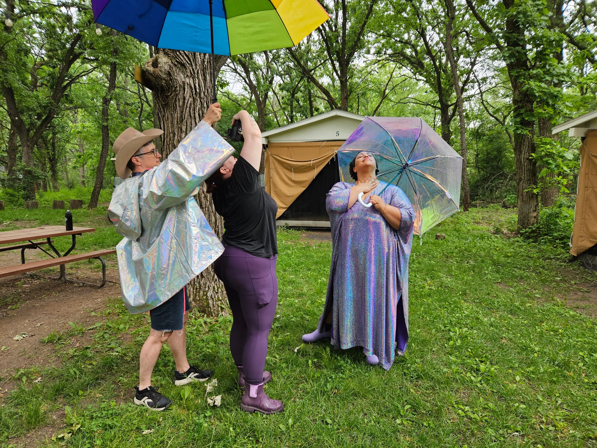 Emily holds a rainbow umbrella over a woman taking photos of another woman who is dressed in an iridescent shawl and holding a similarly iridescent umbrella. In the background, platform tents are visible. They are in a wooded campsite.