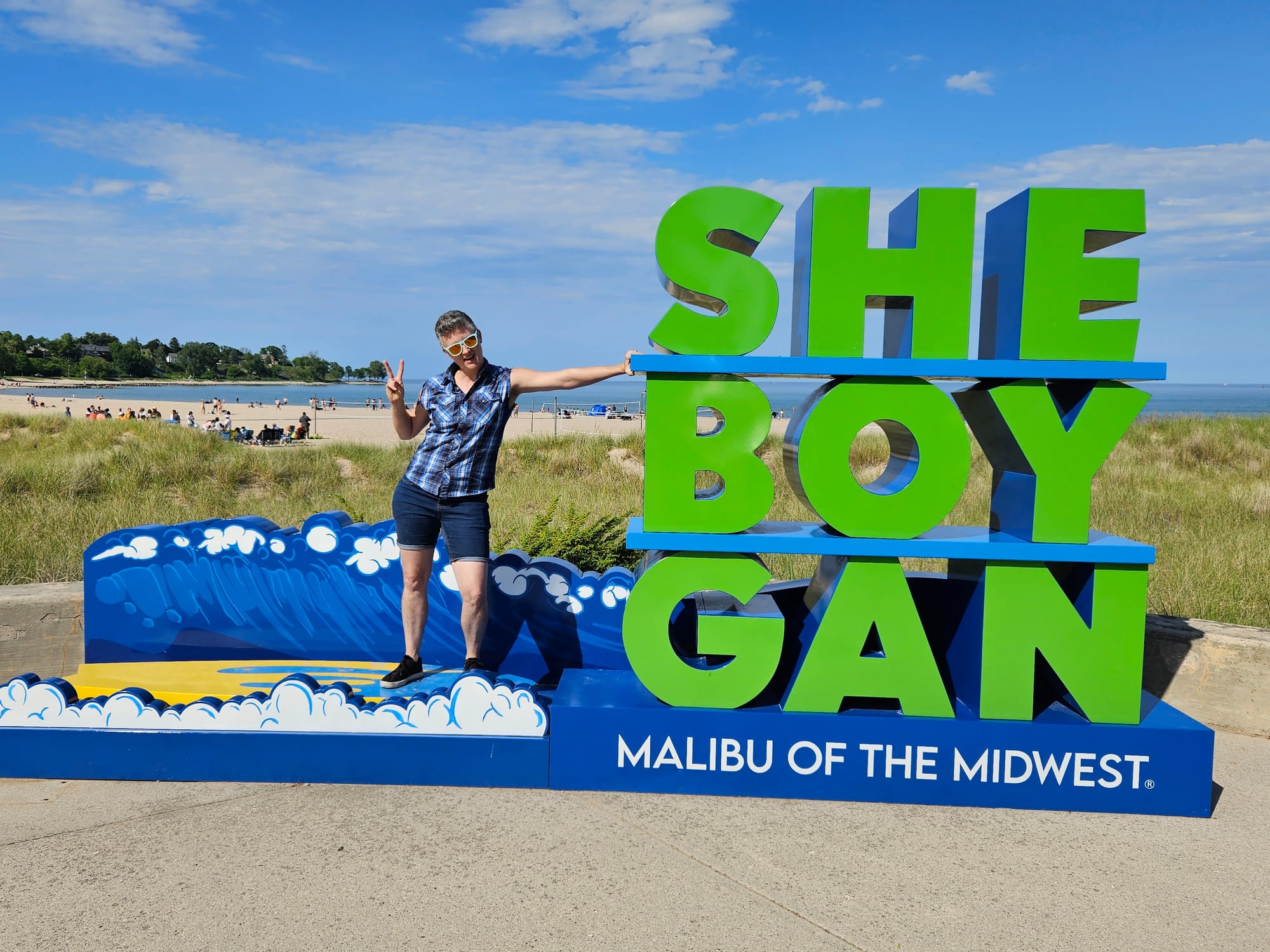 Emily gives the peace sign and poses next to a very large sign that is “SHEBOYGAN” spelled out in big green letters but broken up into SHE, BOY, and GAN, with “Malibu of the Midwest” written underneath.