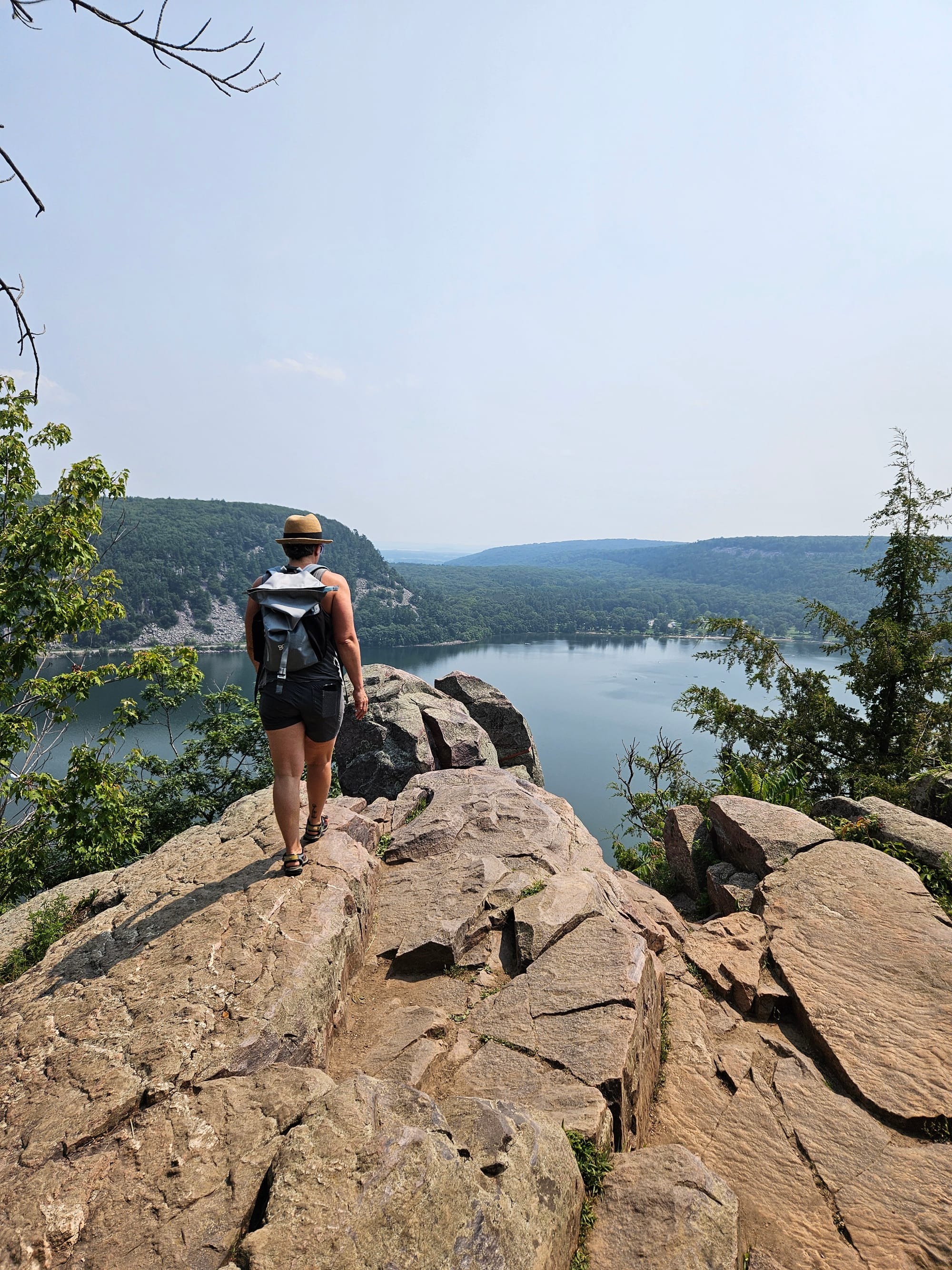 A person walks across rocks on a cliff overlooking a lake and a tree-covered landscape. Haze is visible on the horizon.