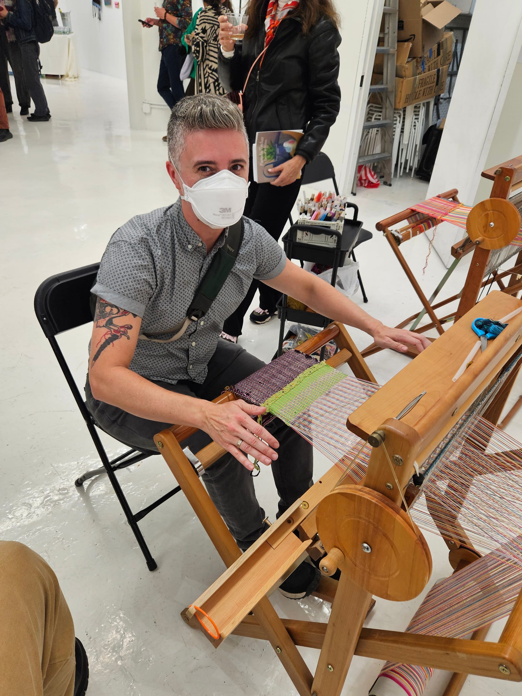 Emily is wearing a KN95 mask and sitting in a chair in front of a large wooden loom. Multi-colored threads are running through it.