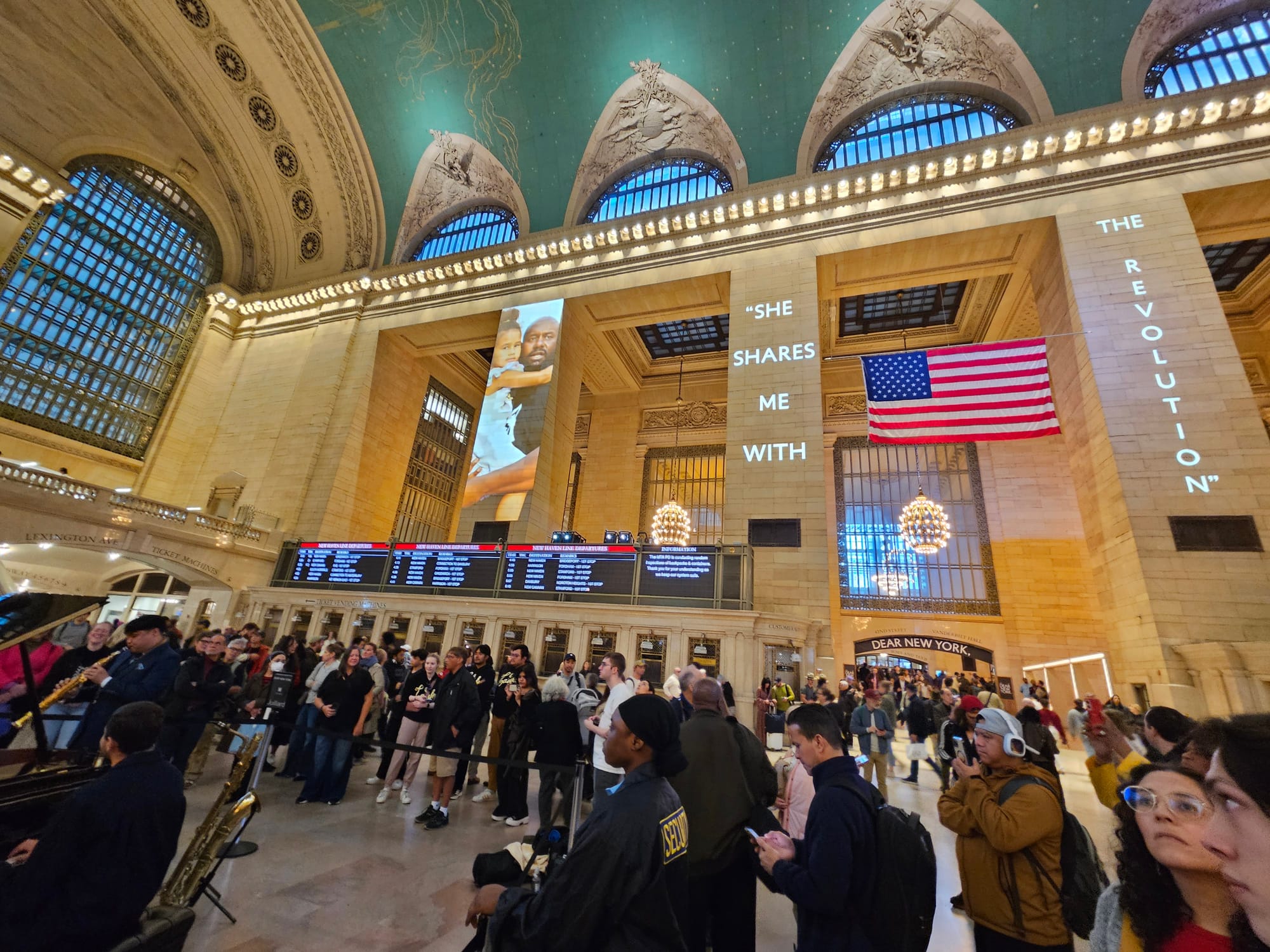 Grand Central’s main gallery is filled with people, many of whom are standing watching musicians perform at a piano in the center. On the marble columns, the projected image of a Black father holding his daughter is paired with the words, “She shares me with the revolution."