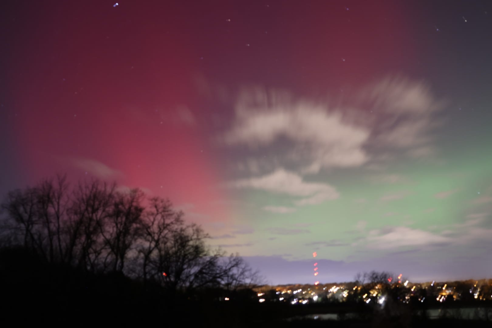 Red and green light streaks the clouds and starry nighttime sky above a line of trees and city lights.