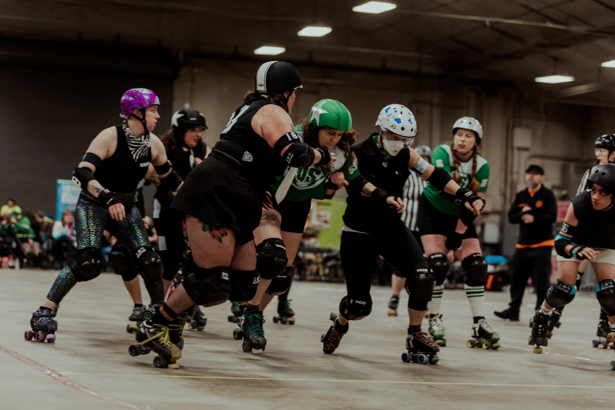 A group of athletes wearing roller skates and protective padding and helmets jostles with each other on an indoor track.