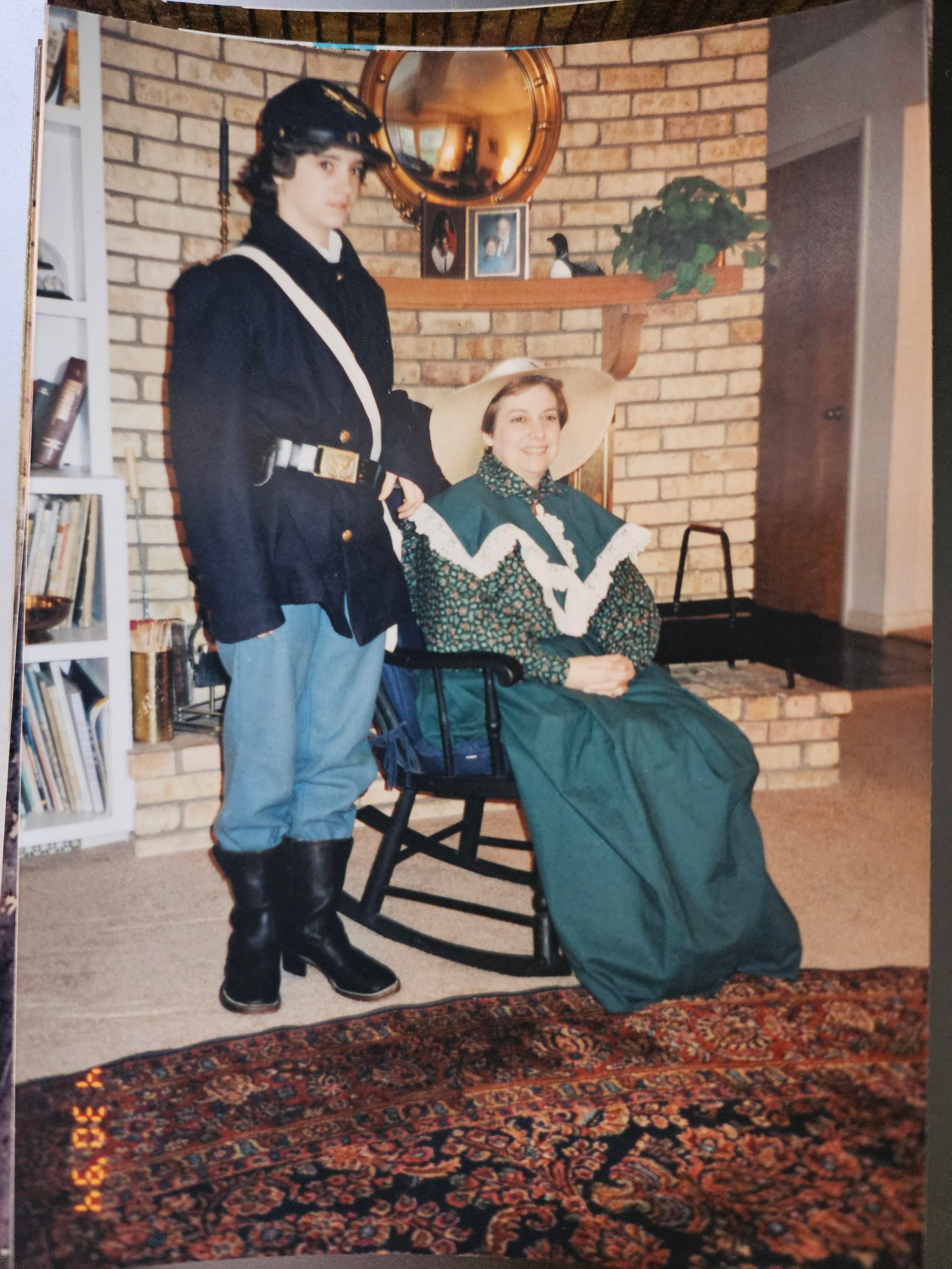 A very young Emily poses in a blue wool Union uniform, standing alongside mom, who sits in a rocking chair and wears an old-timey green dress and big floppy straw hat.