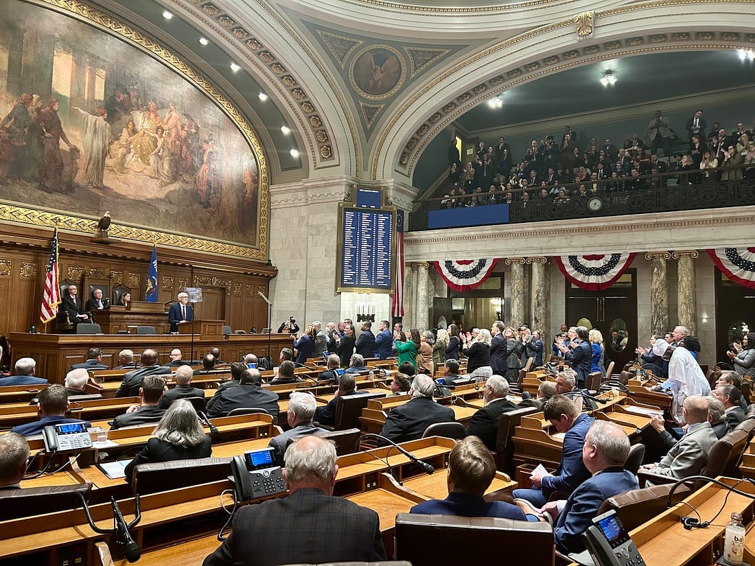 A wide shot of the Wisconsin Assembly Chambers, with members seated at wooden desks and a balcony gallery filled with guests. Gov. Evers stands at a podium at the front of the room.