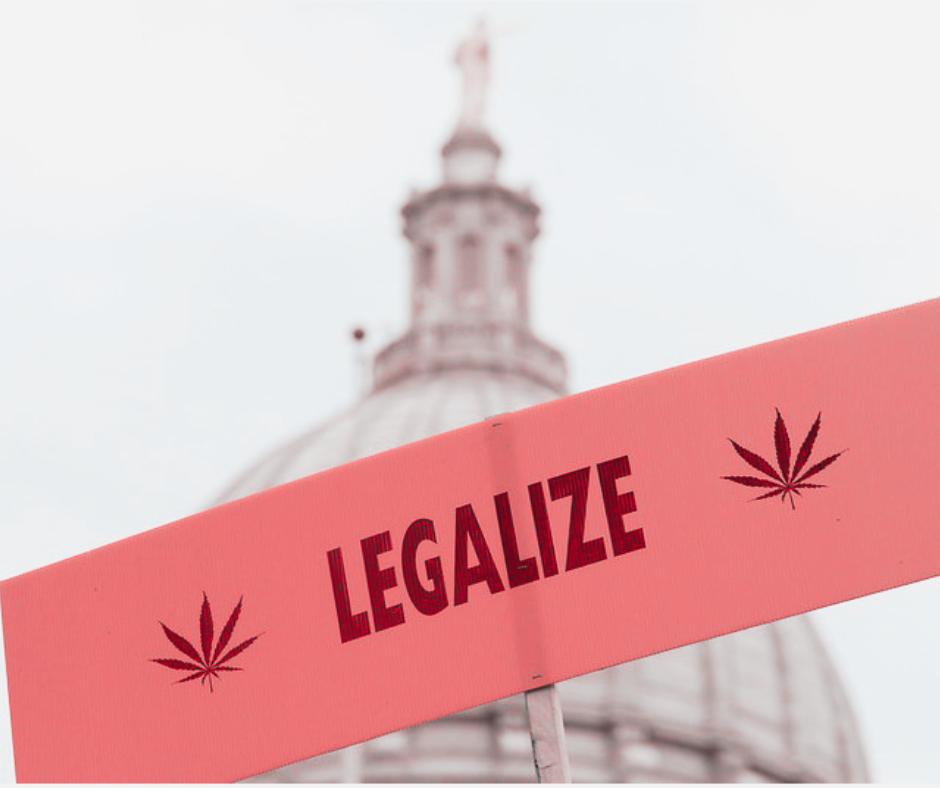 A sign that reads "LEGALIZE" and has illustrations of pot leaves on either side is hoisted in the air in front of the Wisconsin State Capitol building dome.