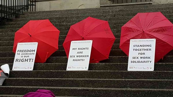 Three red umbrellas with signs leaned against them read: “Freedom to associate is our right!” “My rights are sex worker rights!” “Standing together for sex work solidarity."
