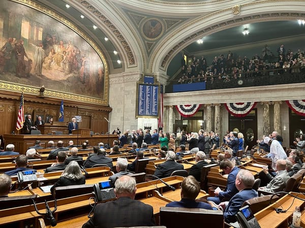 A wide shot of the Wisconsin Assembly Chambers, with members seated at wooden desks and a balcony gallery filled with guests. Gov. Evers stands at a podium at the front of the room.