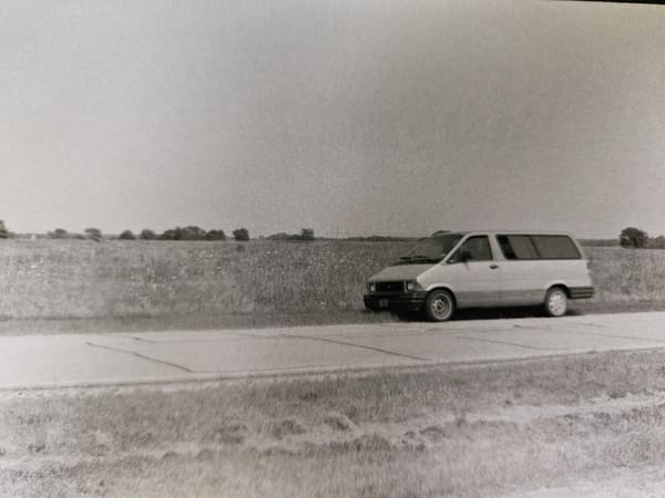 A black and white photo shows an old Ford minivan parked along a cracked roadway, with a flat field of grass and flowers behind it.